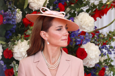 Catherine, Princess of Wales, smiles, dressed in a smart blazer and trousers, with a subtle floral blouse underneath, walking outdoors with a natural background.