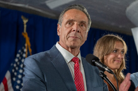 Andrew Cuomo, wearing a suit and tie, stands at a podium with a microphone, gesturing with his hands as he speaks to a crowd.
