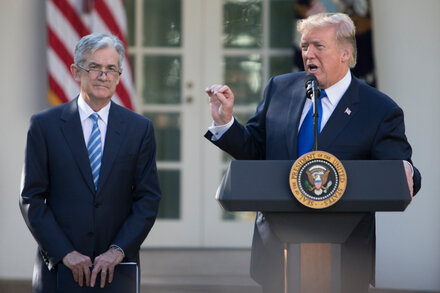 Former President Donald Trump looking at his phone with a serious expression, while Federal Reserve Chair Jerome Powell, in the background, stands at a podium delivering a speech.