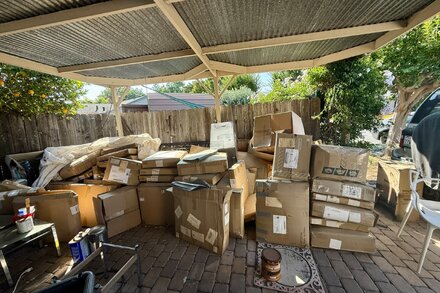 Hundreds of packages are piled high outside a residential home, nearly reaching the roofline. The packages vary in size and are stacked precariously, with some spilling onto the walkway. The scene suggests an overwhelming and unusual accumulation of deliveries.