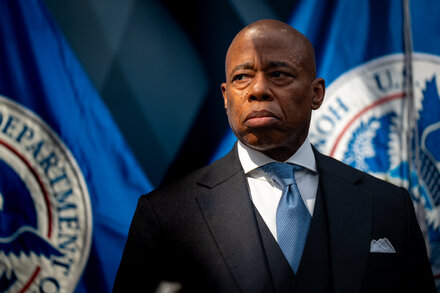 Eric Adams stands behind a podium, looking serious, with a crowd of people in the background. The image likely conveys a sense of political struggle or a challenging moment.