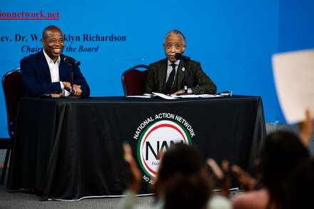 Mayor Eric Adams and Rev. Al Sharpton shake hands at a public event. Adams, a Black man, is wearing a suit and smiling. Sharpton, also a Black man, is wearing a suit and looking at Adams. The background suggests an indoor gathering with other people blurred.