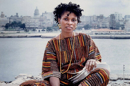 A black and white photo of Assata Shakur, a woman with an afro, looking directly at the camera with a serious expression. She is wearing a light-colored top. The background is simple and out of focus.