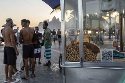 A woman in Brazil uses a digital payment app on her smartphone at a market stall, surrounded by vibrant produce and busy shoppers. The scene highlights the country's embrace of new digital spending habits.