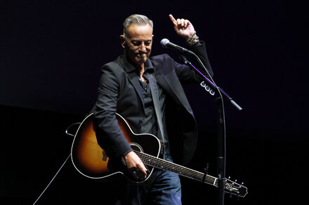 Bruce Springsteen speaks at a microphone on stage, bathed in stage lights, with a serious expression.