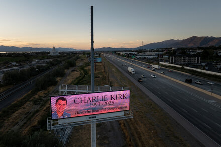 Attendees at a memorial service for Charlie Kirk in Arizona, with a podium and floral arrangements in the foreground and a large American flag in the background.