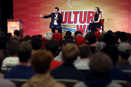 Charlie Kirk speaking at a podium with a serious expression, surrounded by a crowd.