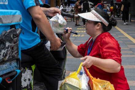 A delivery worker on a scooter navigating through a bustling Chinese city street with tall buildings in the background, possibly carrying packages for a skyscraper delivery.