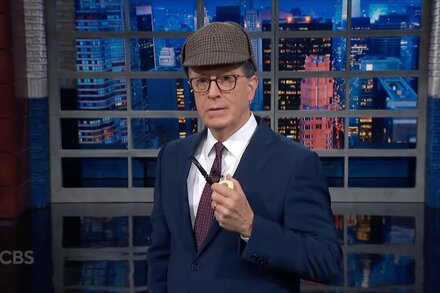 Stephen Colbert looking intently at a screen showing Donald Trump on an escalator, with a questioning expression. The image is a still from The Late Show with Stephen Colbert.