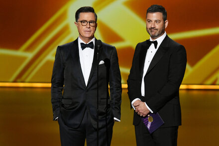 Jimmy Kimmel stands on a stage with a microphone, gesturing to an unseen audience. He is wearing a dark suit and looks like he is telling a joke.