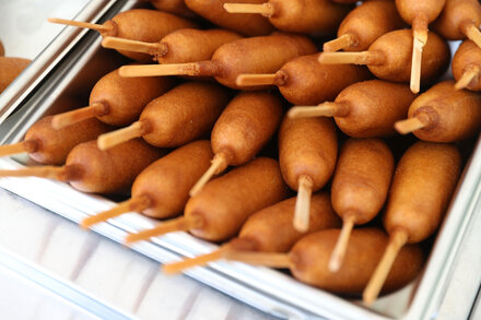 A close-up of a corn dog on a stick, with a bite taken out, showing the sausage inside.