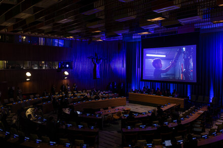 A group of delegates in a large conference room at the United Nations, with the U.N. logo visible. Some delegates are looking at their screens or speaking, indicating a discussion about artificial intelligence.