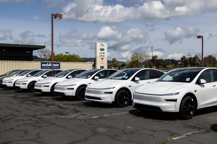 A line of electric vehicles, including sedans and SUVs, sits parked in a dealership lot under a cloudy sky, with a 'SALE' banner visible in the background.