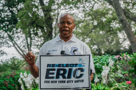 New York City Mayor Eric Adams stands at a podium with a serious expression, gesturing with one hand, against a backdrop of city seals and flags.