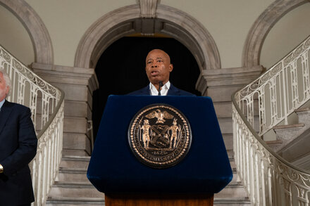 Eric Adams, wearing a suit and tie, stands at a podium with a microphone, gesturing with one hand. Campaign posters are visible in the background, with 'ADAMS' written on them. He appears to be speaking to an audience.