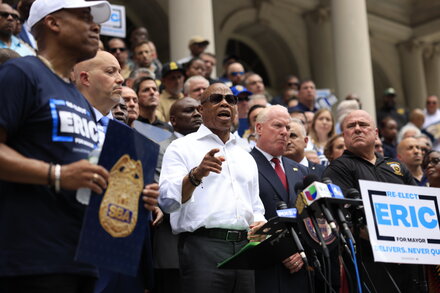 New York City Mayor Eric Adams speaks at a podium, with a timeline of his political career displayed behind him, illustrating key moments and milestones.