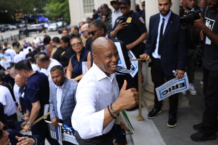 New York City Mayor Eric Adams smiling broadly and raising his arms in a celebratory gesture, wearing a suit and tie.