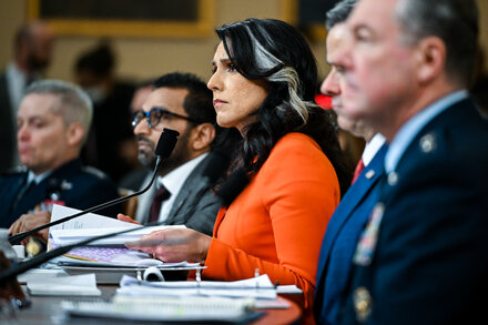 Tulsi Gabbard, a political figure, stands in front of a podium in a formal setting, possibly a press conference or government briefing, with an American flag visible in the background. The image likely depicts her delivering or discussing an intelligence report.