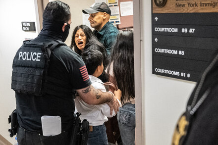 An ICE officer in uniform stands over a woman on the ground, surrounded by onlookers.