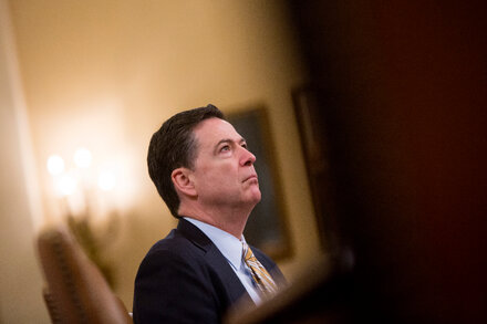Former FBI Director James Comey speaking at a podium with a microphone, looking serious.