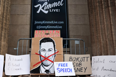 Jimmy Kimmel standing on his talk show stage, holding a microphone and smiling, with the show's logo in the background.