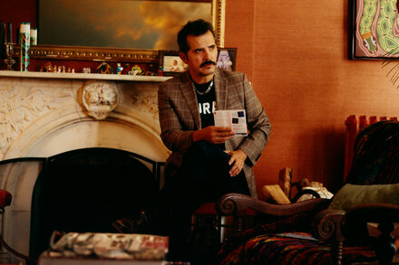 John Leguizamo, a man with short dark hair and a beard, stands in front of a mirror in a dressing room, applying makeup. He is wearing a dark t-shirt and looks focused. Various makeup brushes and containers are visible on the counter in front of him.