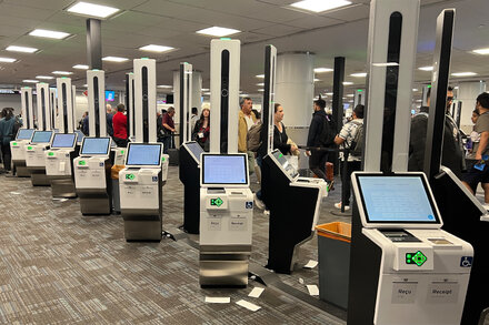 Passengers queue at check-in counters with airport self-service kiosks showing error messages or being unused due to a system outage at a busy Canadian airport.
