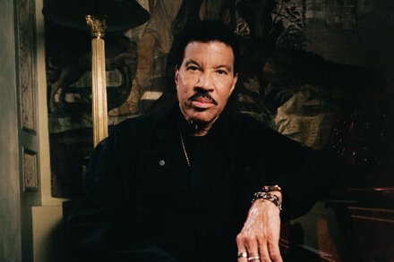 Lionel Richie, wearing a black shirt and a serious expression, stands with his arms crossed, looking directly at the camera.
