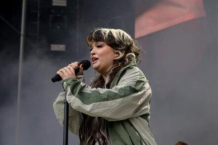 Lola Young performs on stage, holding a microphone and looking visibly distressed, with a band in the background.