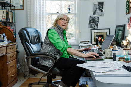 Marilyn Hagerty smiling warmly, holding a pen and notepad, with a backdrop of a restaurant interior. She is an elderly woman with short, styled white hair, wearing glasses and a patterned top.