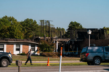 Investigators examining a damaged church in Michigan where a shooting occurred, with police tape visible and emergency vehicles in the background.