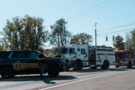 A police car with flashing lights blocks a road near a church. Smoke rises from the church building in the background.