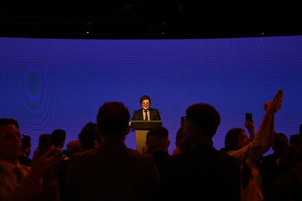 Javier Milei, Argentina's president, standing in front of a blue backdrop, delivering a speech. He has a serious expression, and his hand is raised, possibly in a gesture related to his economic policies.