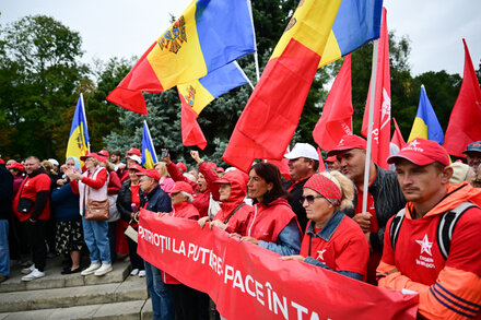 A group of people, some holding Moldovan flags, gather in a public square. The architecture suggests a European city. Some appear to be listening to a speaker or watching an event.