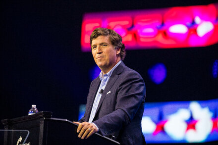 A man speaks into a microphone on a stage with a screen behind him showing a red and blue graphic.