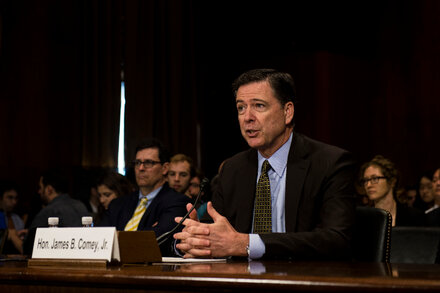 James Comey, former FBI director, testifies before a congressional committee. The image also subtly references a norm-shattering indictment and Amazon's $2.5 billion settlement.