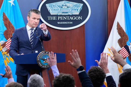A reporter holding a microphone speaks to a Pentagon official in a hallway, with other reporters and cameras visible in the background, illustrating the restricted access.