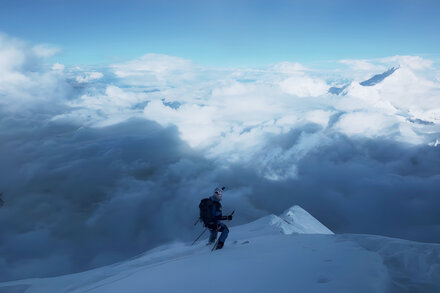 Polish skier Andrzej Bargiel in climbing gear with skis on backpack, looking at Everest summit, with a panoramic view of the Himalayas and clouds below.