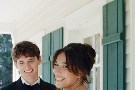 A bride and groom in a dark, elaborately decorated room that looks like a haunted house, with cobwebs and vintage furniture. The bride is in a white dress with a black veil, and the groom is in a dark suit.