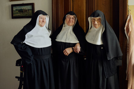 A group of nuns, some smiling, stand together in a sunlit courtyard, with a historic European building in the background. They appear to be in conversation or posing for a photo.