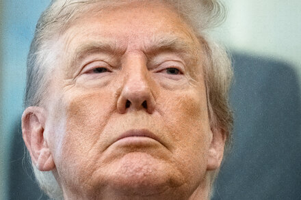 Donald Trump in a suit and red tie, standing at a podium with a microphone, gesturing with his hand. The background is a blurry American flag.