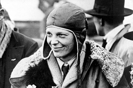 President Trump behind a podium, gesturing, with an image of Amelia Earhart and her plane in the background, overlaid with text 'Amelia Earhart: Unsealed Files'