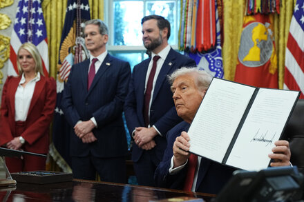 Donald Trump seated at a desk, signing a document, with a microphone and an American flag in the background, likely related to the TikTok deal.