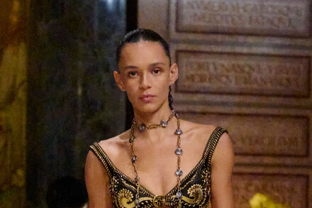 Models walk down a muddy runway at a Versace fashion show, showcasing garments stained with dirt and grime, reflecting a rebellious and raw aesthetic.