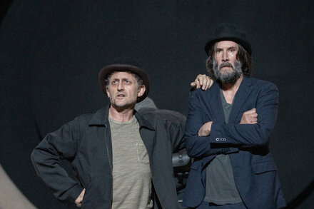 Actors on stage in a 'Waiting for Godot' production, one possibly holding an air guitar or striking a theatrical pose.