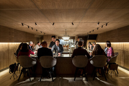 A chef meticulously plates a gourmet dish in a high-end restaurant kitchen, with blurred background hinting at a busy but elegant dining experience. The focus is on the intricate presentation of food, suggesting culinary excellence.