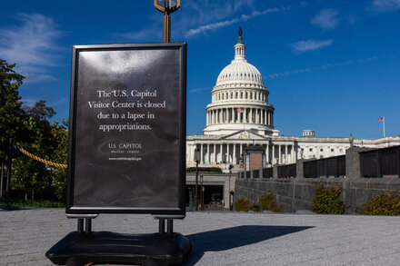 A woman in a suit walks past a federal building with a sign indicating it is closed due to a government shutdown. She looks thoughtful.