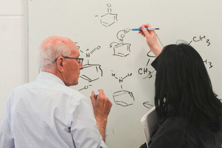 Three scientists with serious expressions are seated in front of a chalkboard filled with complex equations and scientific diagrams. One scientist, in the center, gestures with a pen while looking at a beaker filled with colorful liquids. The other two scientists are looking intently at a microscope.