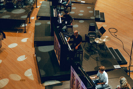 Fifty grand pianos arranged in a large performance hall, with a few musicians visible. The image captures the scale and grandeur of the '11,000 Strings' concert.