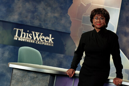Belva Davis smiles, looking directly at the camera. She wears a dark suit jacket over a light blouse, and a brooch is pinned to her lapel. Her dark hair is styled short.
